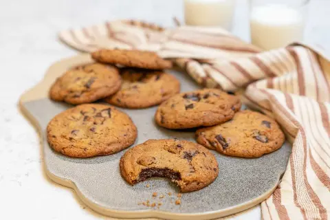 Nybakade chocolate chip cookies recept på en grå platta, med en bit tagen ur en cookie. Två glas mjölk och en randig handduk i bakgrunden.