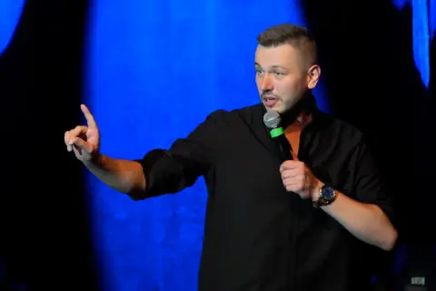 Mężczyzna w czarnej koszuli, trzymający mikrofon, gestykuluje na scenie. To jego top stand up.