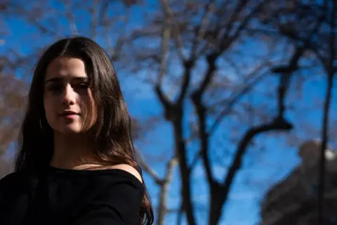 Elvira Lara, una joven con cabello castaño, mira a la cámara con un fondo de árboles y cielo azul.