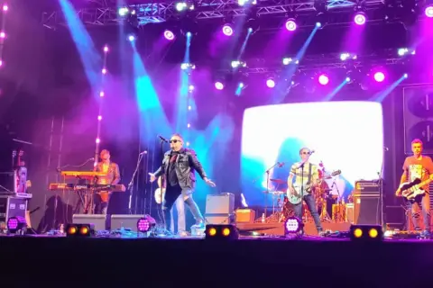 Zespół gra na scenie podczas koncertów w Stalowej Woli. Wokalista w skórzanej kurtce, gitarzyści i klawiszowiec.