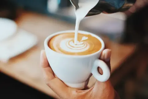 En hand håller en vit kopp med en vacker kaffe latte, där mjölken skapar ett hjärta i skummet.