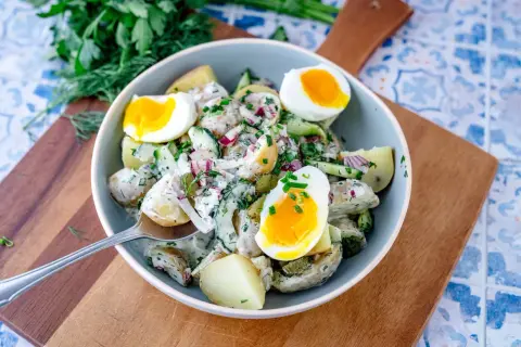 Schüssel mit cremigem Kartoffelsalat mit Gurken, Zwiebeln und gekochten Eiern, garniert mit frischen Kräutern.