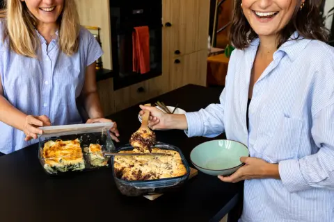 Deux femmes souriantes préparent la conservation de fonds de tarte cuits dans des plats en verre. L'une sert une part d'un plat de tarte au chocolat, l'autre ferme un plat de lasagnes.