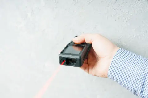 Dłoń trzyma dalmierz laserowy, z którego wychodzi czerwona wiązka. To najlepszy laser do precyzyjnych pomiarów.