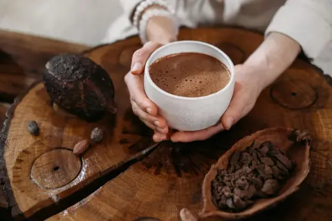 Hände halten eine Tasse heißen Kakao, umgeben von Kakaobohnen und einer Kakaoschote. Ein Moment der Ruhe bei einer Kakao-Zeremonie.