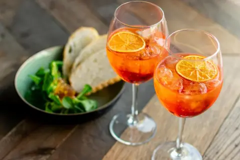 Zwei Gläser mit einem orangen Aperitif, garniert mit Orangenscheiben, daneben ein Schälchen mit Salat und Brot.