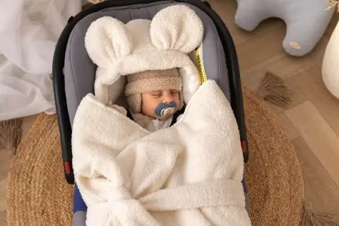 Słodki maluszek śpi w miękkim, kremowym otulaczu do fotelika z uszkami.