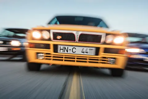 Żółta Lancia Delta Integrale w centrum, ścigana przez niebieski i czarny samochód.