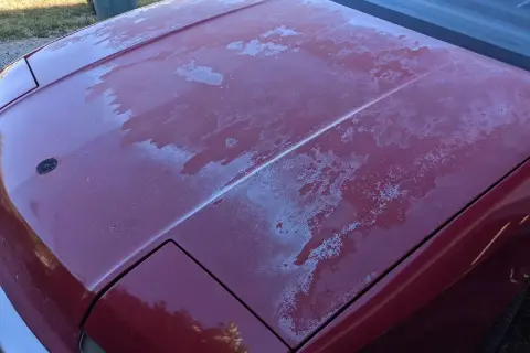 Il cofano di un'auto rossa presenta il trasparente rovinato, con chiazze bianche che indicano la necessità di capire come togliere il trasparente dalla carrozzeria.