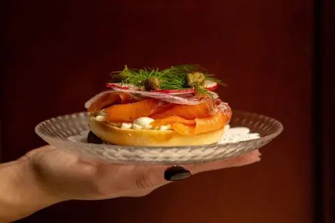 En hand håller upp en bagel med rökt lax, färskost, rödlök, rädisor och kapris. Perfekt för en lyxig bank hotel middag.