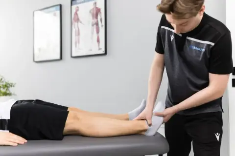 Fizjoterapeuta w sporcie delikatnie bada stopę zawodnika leżącego na kozetce.
