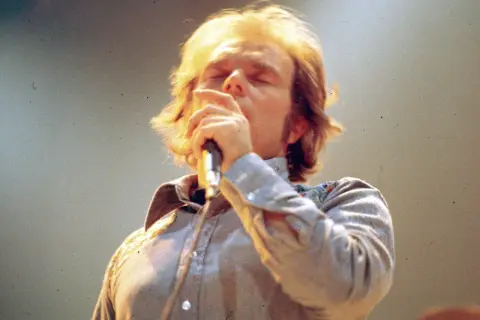 Van Morrison sings passionately into a microphone, his eyes closed. This image captures a moment from one of his legendary performances, perhaps hinting at the setlists that have defined his career.