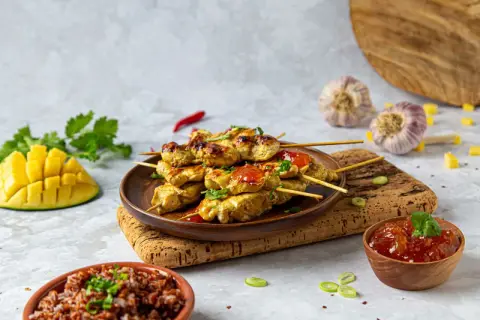 Aksamitne satay z kurczaka na patyczkach, polane sosem, obok ryż, mango i czosnek.