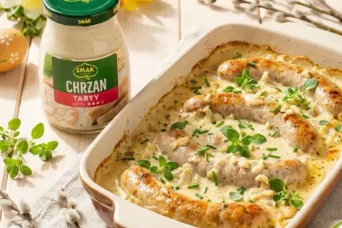 Biała kiełbasa w sosie chrzanowym, zapiekana w naczyniu, z dodatkiem ziół. Obok słoik sosu chrzanowego.