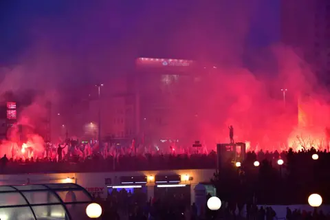 Tłum z polskimi flagami, race i dym tworzą atmosferę manifestacji. W tle widać budynki i billboard Santander. To obraz silnego nacjonalizmu.