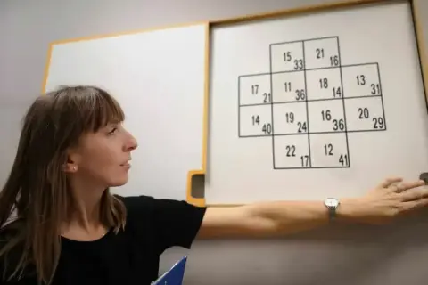 Kobieta przeprowadza testy psychologiczne dla kierowców, wskazując na tablicę z liczbami.