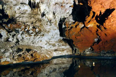 Interior de una cueva con estalactitas y formaciones rocosas rojizas, reflejándose en un espejo de agua. El **cerro del hierro** se revela en su esplendor subterráneo.