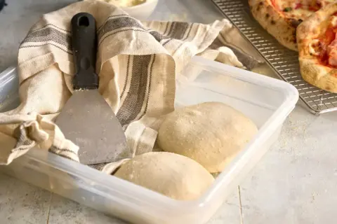 Pizzadeg redo att baka ut. En degskrapa och en kökshandduk ligger bredvid en plastlåda med två degbollar.