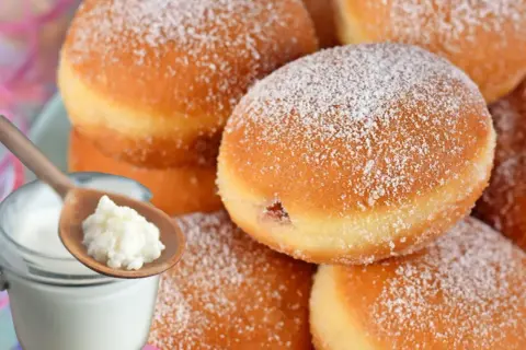 Puszyste pączki z lukrem, posypane cukrem pudrem, obok słoiczka z twarożkiem i drewnianej łyżeczki.