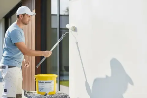 Mann streicht mit einer Fassadenrolle eine weiße Wand. Ein Eimer mit Farbe steht daneben.