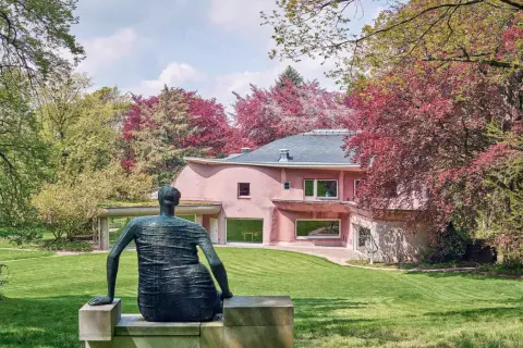 Bronze-Figur blickt auf rosafarbenes Haus im Skulpturenpark Waldfrieden, umgeben von leuchtend rotem und grünem Laub.