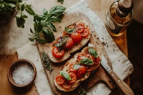 Två bitar bruschetta med mozzarella, färska tomater och basilika på en träskärbräda. En flaska olivolja och en skål med flingsalt syns bredvid.