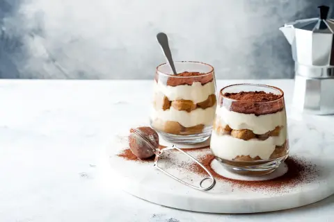 Dwa pucharki z tiramisu bez jajek, przełożone biszkoptami i kremem, posypane kakao. Obok sitko do kakao i kawiarka.