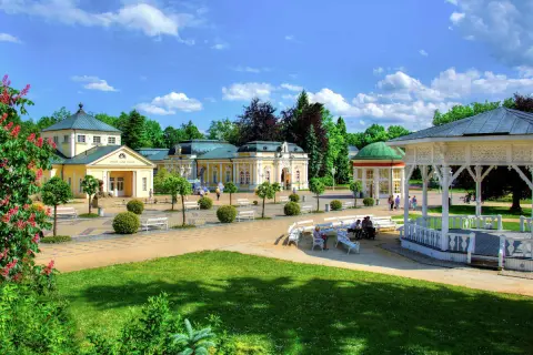 Ein sonniger Tag in einer **Therme Tschechien**. Gelbe Kurgebäude mit grünen Dächern, ein weißer Pavillon und gepflegte Gärten laden zum Entspannen ein.