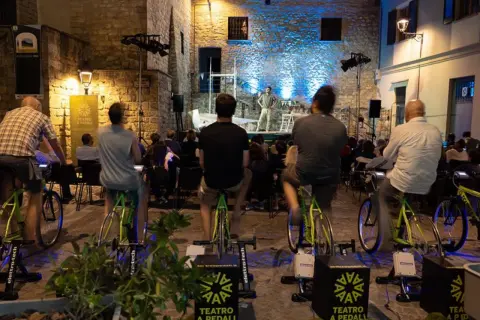 Spettatori pedalano su cyclette per alimentare un'esibizione di teatro a pedali in una piazza illuminata.