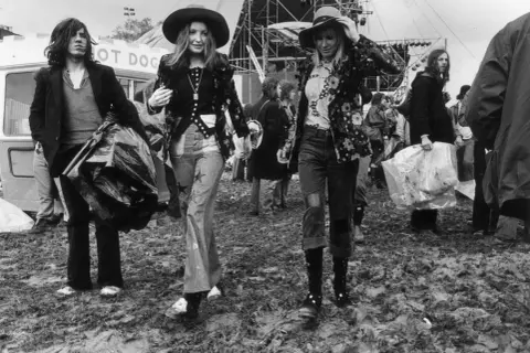 Moda hipisowska lat 70. na festiwalu. Dwie kobiety w kapeluszach i kwiecistych kurtkach, mężczyzna z torbą.