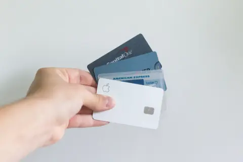 Dłoń trzyma kilka kart płatniczych, w tym Apple Card, American Express i Capital One. Standardowe wymiary karty bankomatowej są widoczne.
