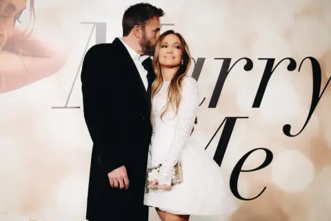 Ben Affleck całuje w czoło Jennifer Lopez, która w białej sukni trzyma torebkę. Para wygląda na zakochanych.