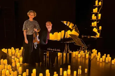 Músicos disfrutan de un concierto con velas. Una violinista y un pianista posan junto a un piano rodeado de velas encendidas.
