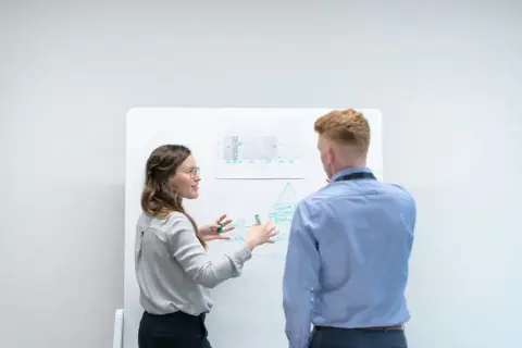 Kvinna förklarar en modell på whiteboarden för en man. Diskussionen kan handla om skillnad mellan befattningsbeskrivning och arbetsbeskrivning.