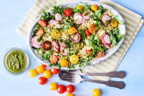 En stor bulgursallad med körsbärstomater, rädisor och ruccola, toppad med en grön dressing. Serveras med en skål pesto och extra tomater.