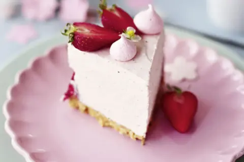 Ein Stück Erdbeer Cheesecake mit frischen Erdbeeren und Baiser-Tupfern auf einem rosa Teller.