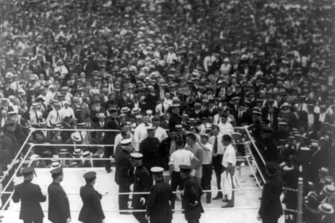 Boxing ring with fighters and officials, surrounded by a massive, cheering crowd in vintage attire.