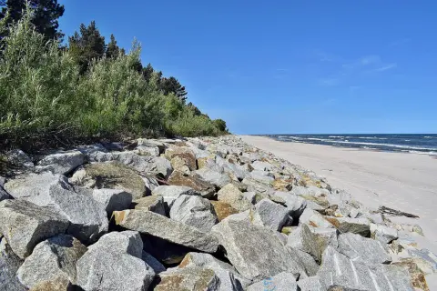 Kamienisty brzeg morza z zielenią i piaszczystą plażą w tle.