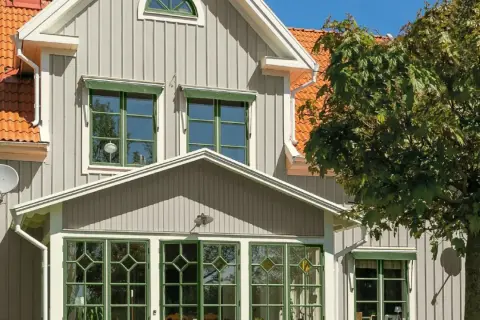 Vitt hus med gråa knutar och orange tak. Gröna fönster och en stor lönn skänker skugga.