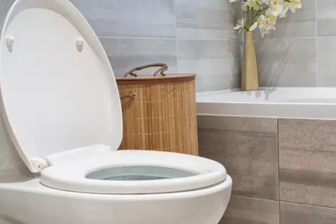 Biała deska WC z otwartą klapą, obok bambusowy kosz i wazon z kwiatami. Dowiedz się, jak dobrać deskę WC do swojej łazienki.