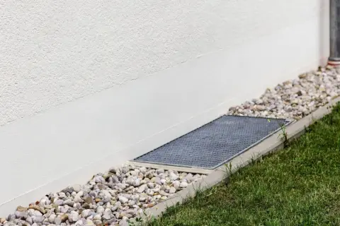 A gravel bed and grate next to a white wall, showing how to level ground for drainage.