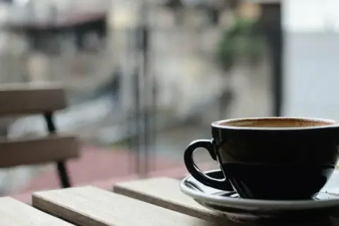 En svart kopp med lungo kaffe står på ett träbord. I bakgrunden syns en bänk och en suddig stadsmiljö.