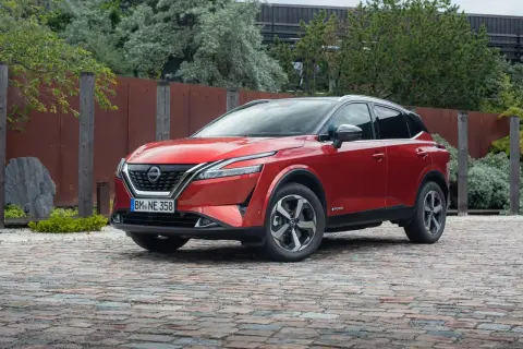 Czerwony Nissan Qashqai 2025 z czarnym dachem, zaparkowany na brukowanej nawierzchni, otoczony zielenią i industrialnym ogrodzeniem.