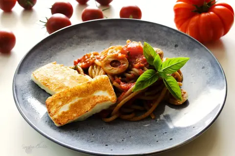 Makaron z halloumi na talerzu, z sosem pomidorowym, oliwkami i bazylią. Obok leżą świeże pomidory.