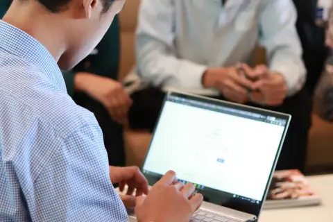 Mężczyzna w niebieskiej koszuli pracuje na laptopie. W tle widać innych ludzi, być może to spotkanie z kontrahentem.
