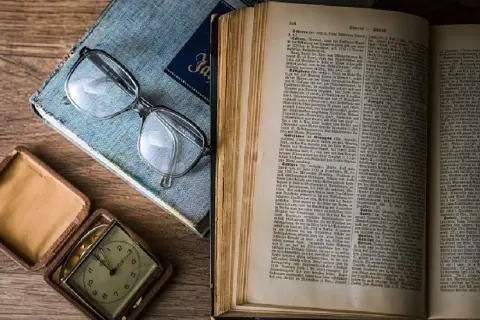 Okulary leżą na niebieskiej książce, obok otwarte pudełko z zegarkiem i otwarta księga, jakby czytano Eneidę.