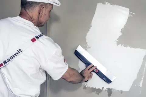 Fachowiec w białej czapce i koszulce z logo Baumit wykonuje gipsowanie ścian, używając szerokiej pacy do rozprowadzania masy.
