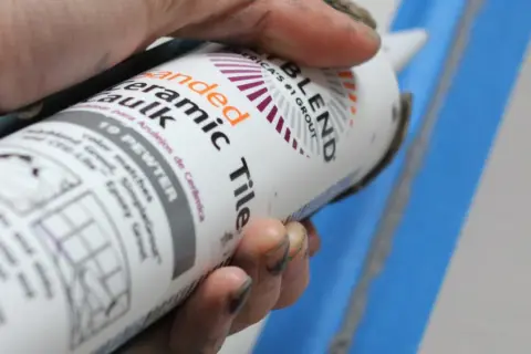 Hand holding a tube of "Sanded Ceramic Caulk" in "19 Pewter" color.