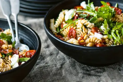 Zwei Schalen mit einem köstlichen nudelsalat mit getrockneten tomaten, Pinienkernen und Rucola, garniert mit Feta.