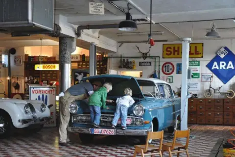 Familie bei der Oldtimer-Restauration eines blauen Opels. Der Vater erklärt den Kindern den Motorraum.